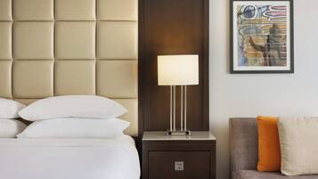 Una camera d'albergo con una testiera imbottita, un comodino con una lampada, un quadro astratto incorniciato e un divano con cuscini.
