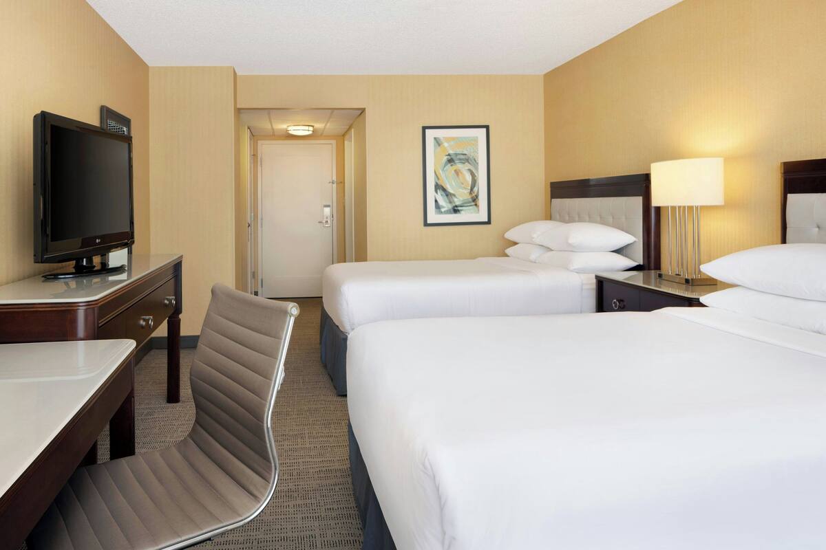 A hotel room with a bed, bedside table, lamp, TV, chair, and artwork on the wall.