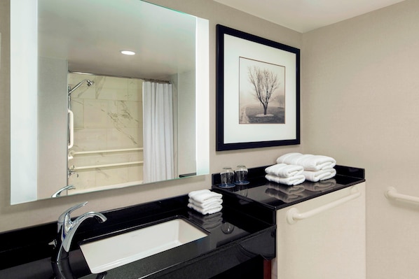 A modern bathroom with a black countertop, a white sink, a large mirror, a framed picture of a tree, and a shower area. - Sheraton West Des Moines Hotel (West Des Moines)