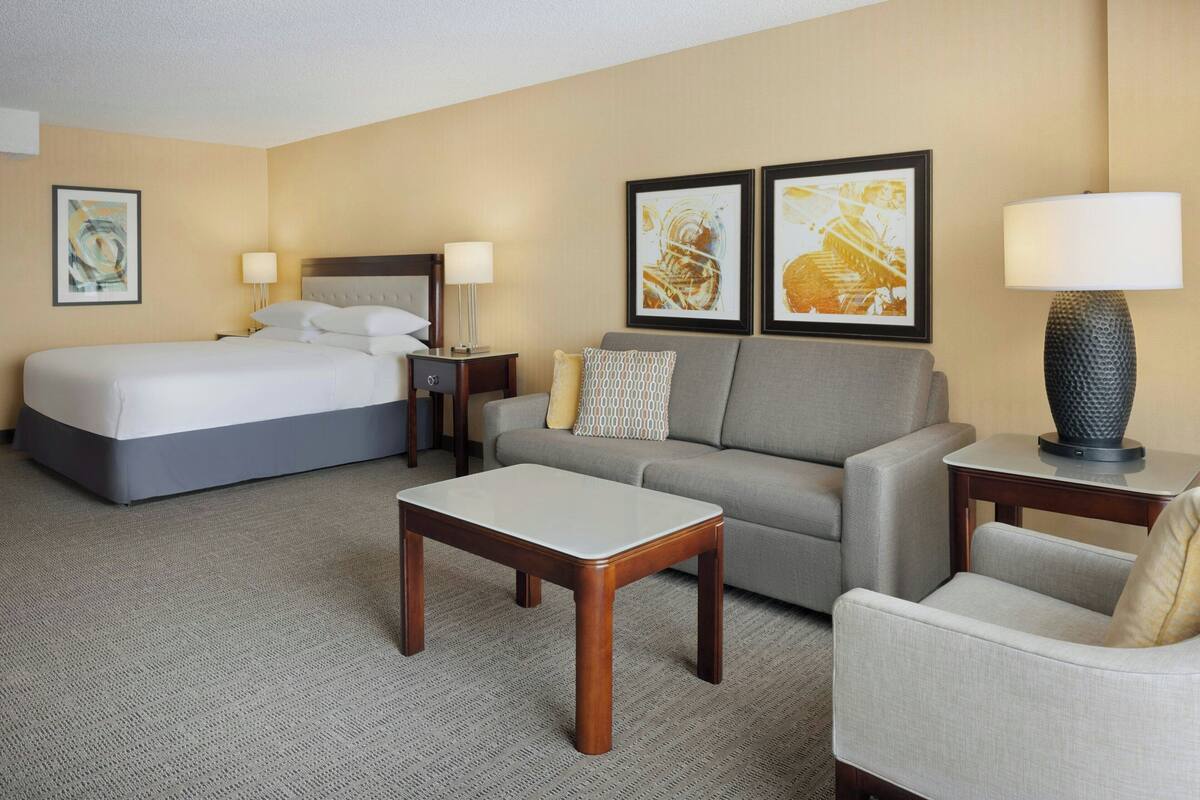 A hotel room with a bed, a sofa, a coffee table, a chair, and two framed artworks on the wall.