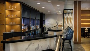 Una zona de bar con mostrador de mármol, estantes con copas y botellas, y un área de descanso con sillones.