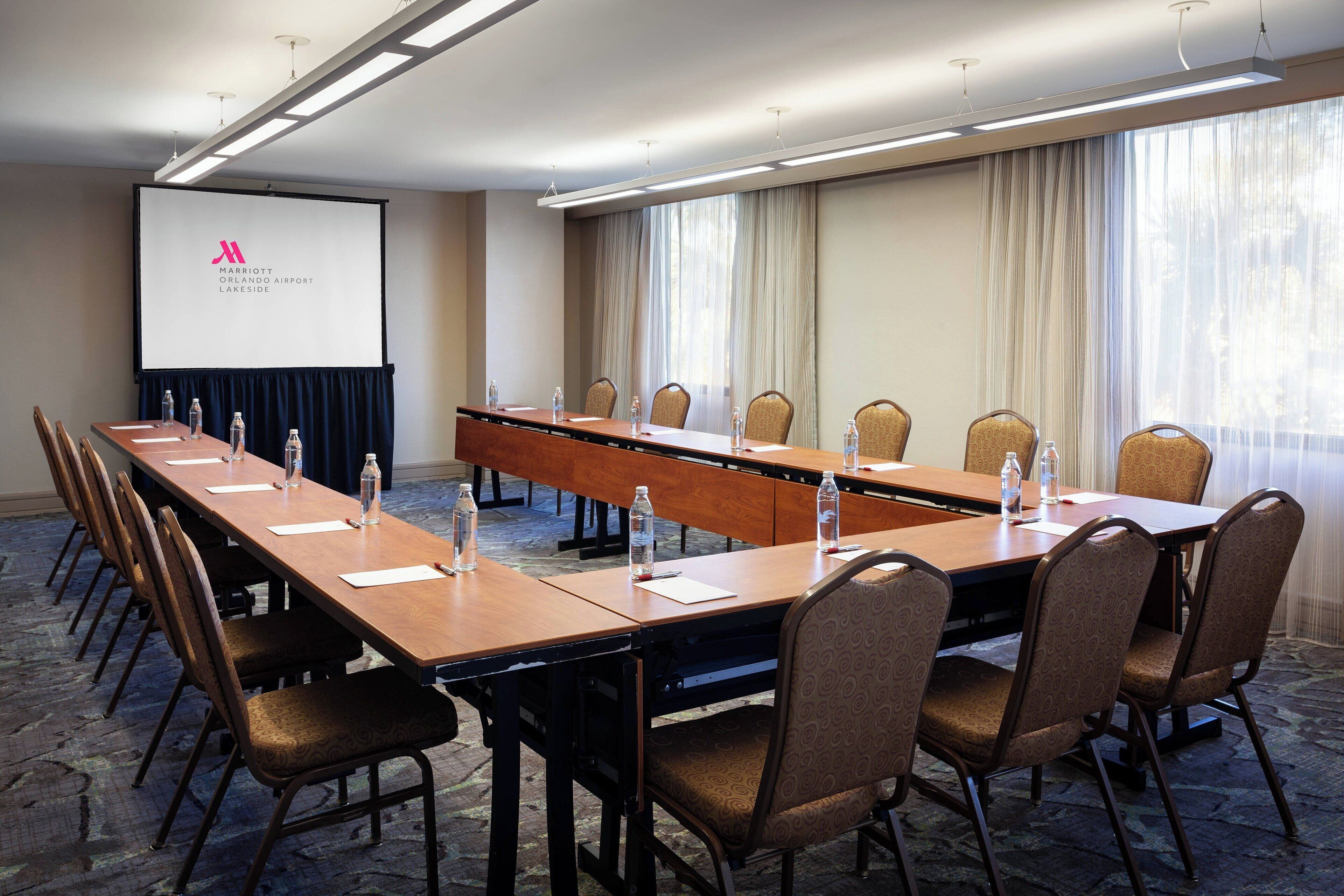 A conference room with a long table, chairs, a projector screen, and a large window with curtains.