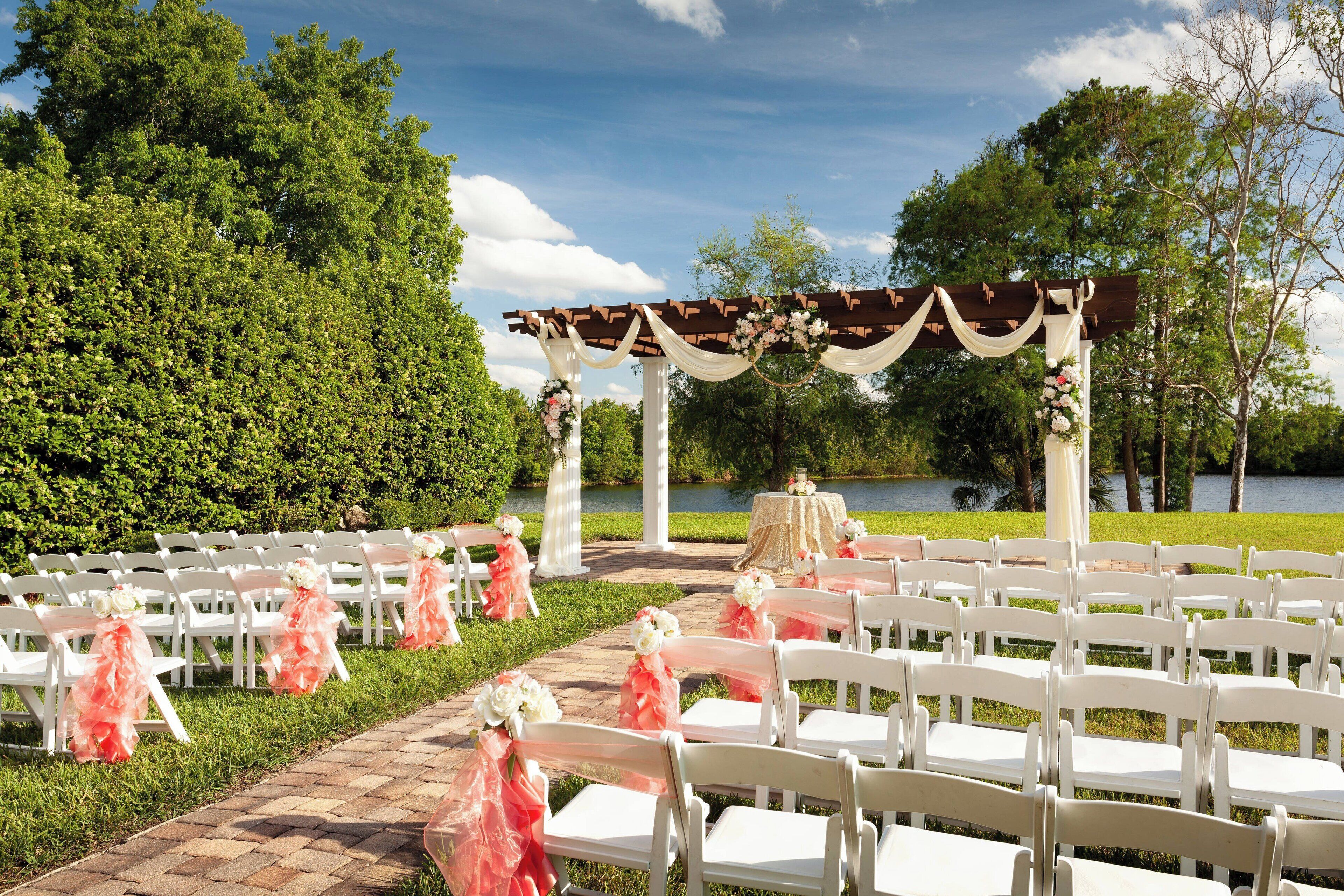 An outdoor wedding setup with white chairs arranged in rows, a wooden pergola with draped fabric, and a table with a white tablecloth.