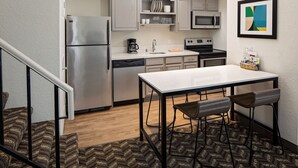 Una cocina compacta equipada con refrigerador, microondas, cafetera y lavavajillas. Hay una mesa de comedor con dos sillas y una alfombra pequeña en el suelo.