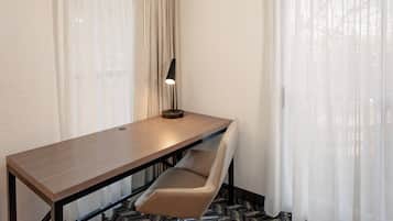 A hotel room with a wooden desk, a modern chair, a lamp, and sheer curtains.