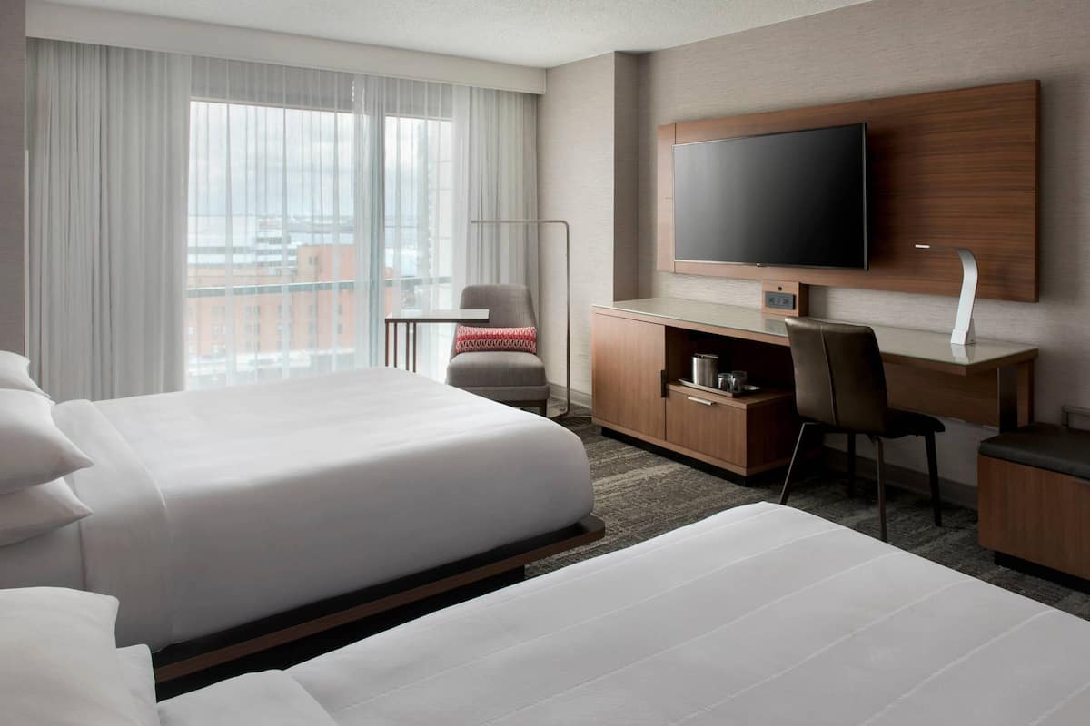 A hotel room with two beds, a desk, a TV, and a view of the city.