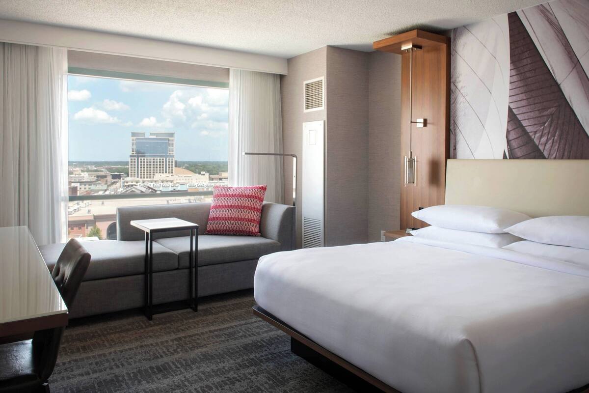 A hotel room with a large bed, a sofa, a small table, and a view of a cityscape through a large window.