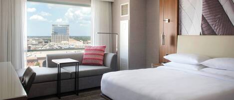 A hotel room with a large bed, a sofa, a small table, and a view of a cityscape through a large window.