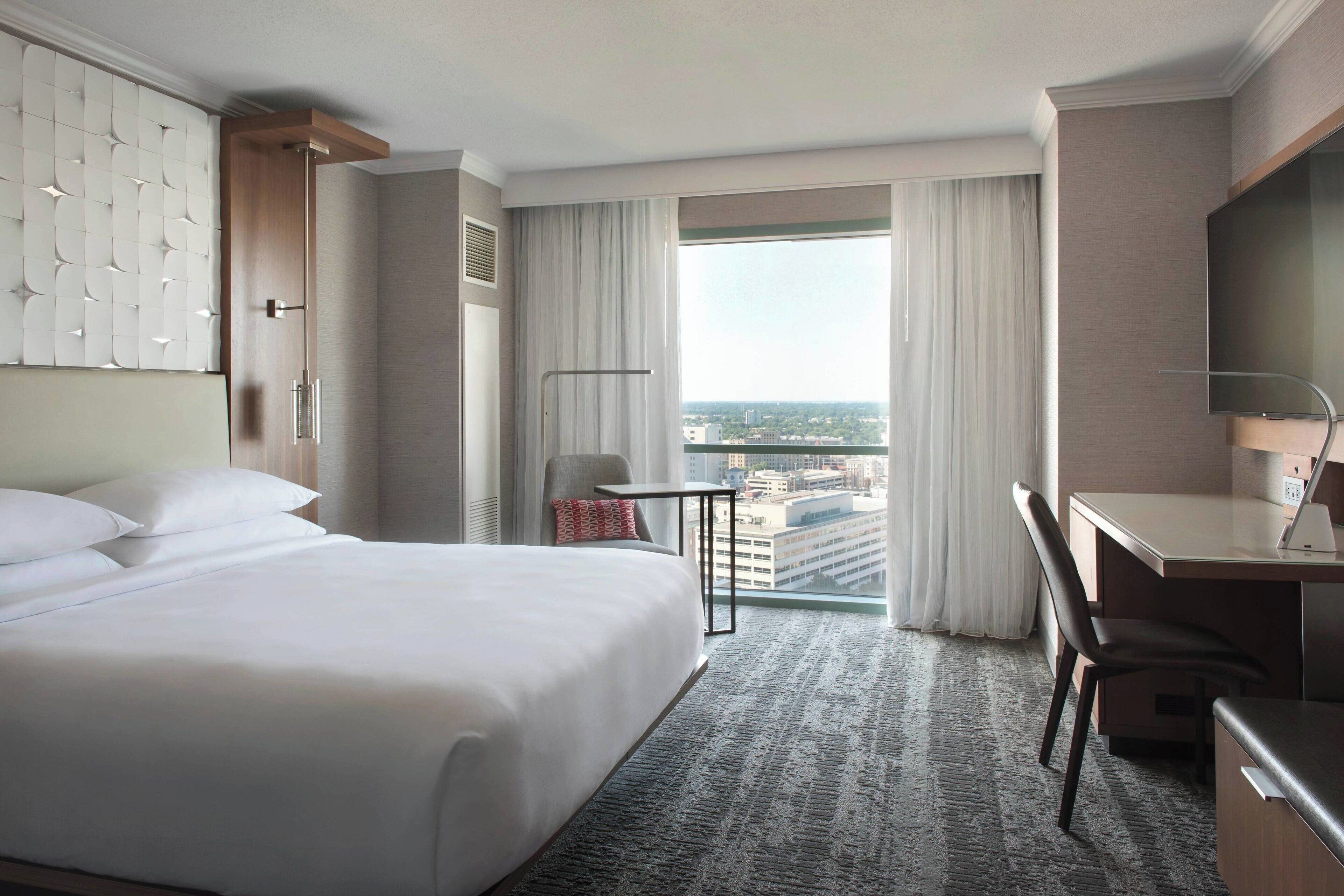 A hotel room with a large bed, a desk, a chair, a small table, and a view of the city through the window.