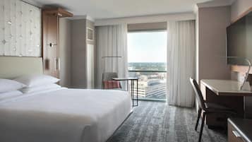 A hotel room with a large bed, a desk, a chair, a small table, and a view of the city through the window.
