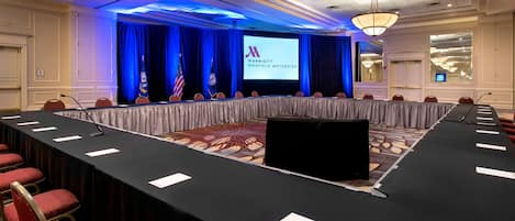 Une salle de conférence avec une longue table, des chaises rouges, des drapeaux et un écran affichant le logo de Marriott.