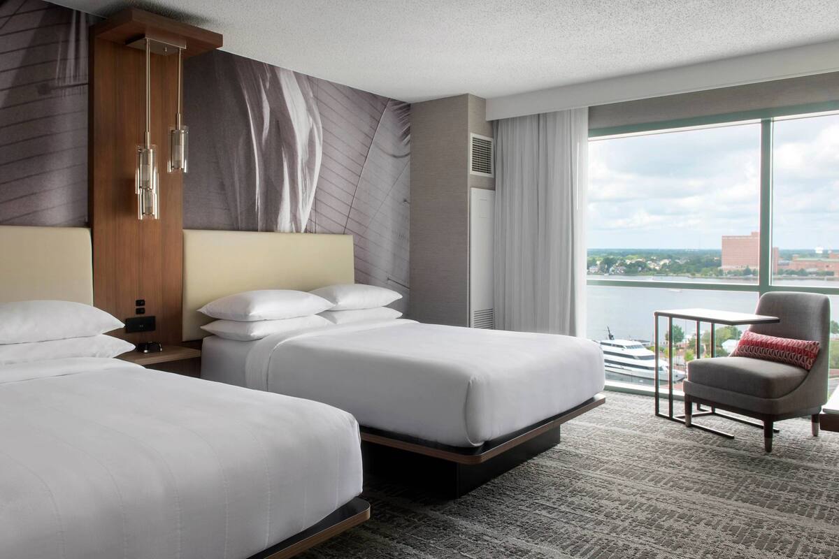 A hotel room with two beds, a large window with a view, a chair, and a small table.