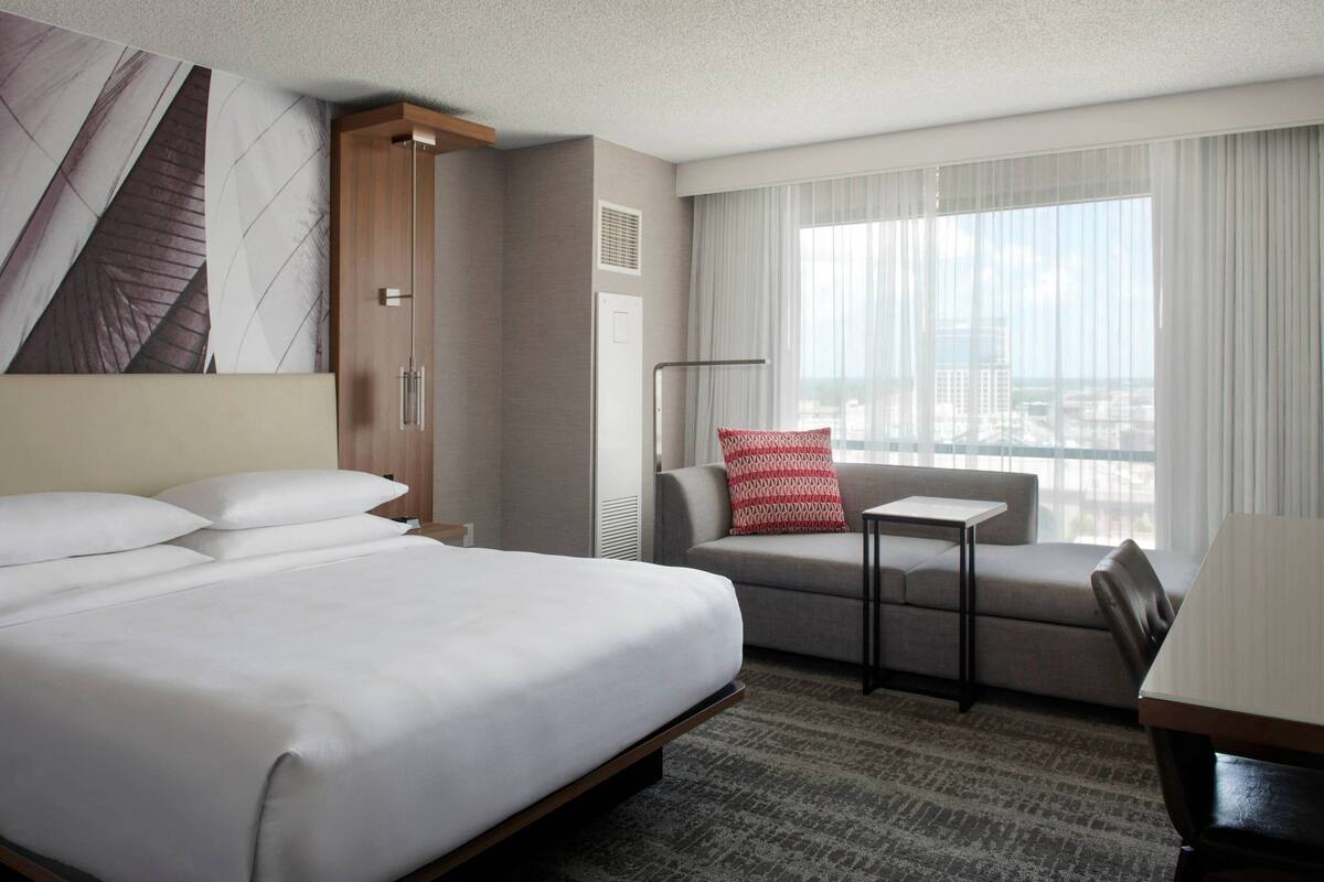 A hotel room with a large bed, a sofa, a small table, and a view of the city through large windows.