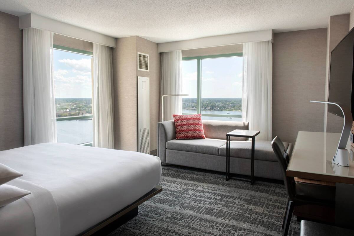 A hotel room with a large bed, a desk, a chair, a small table, and a window with a view of a cityscape.