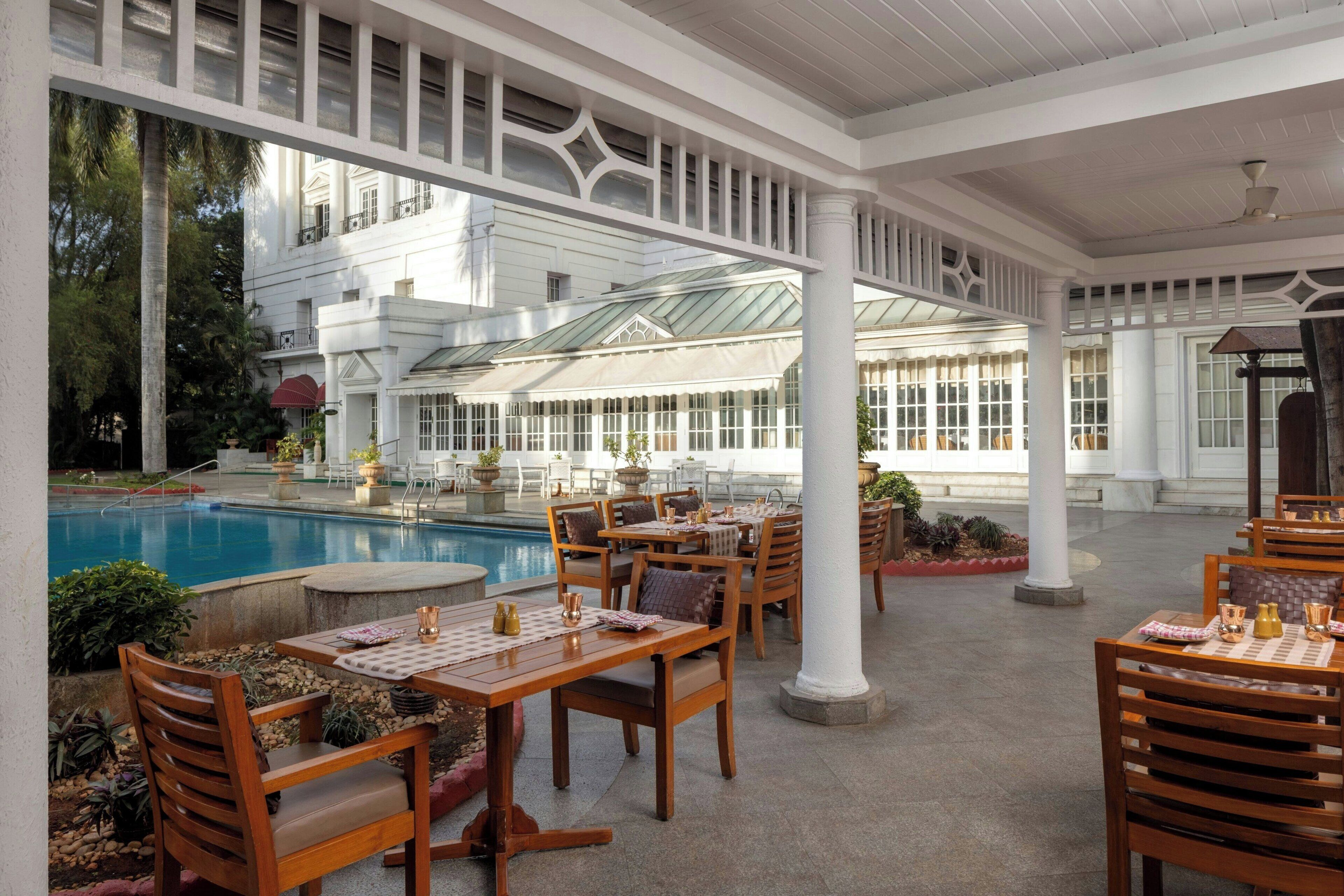 An outdoor patio with wooden tables and chairs, a swimming pool, and a building with large windows in the background.