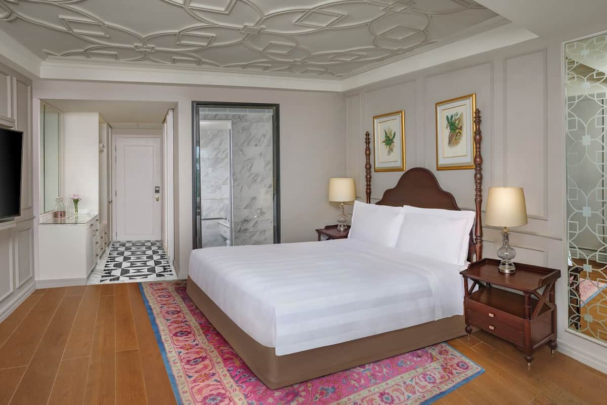 A hotel room with a large bed, two bedside tables with lamps, a television, a bathroom with a shower, and a patterned rug.