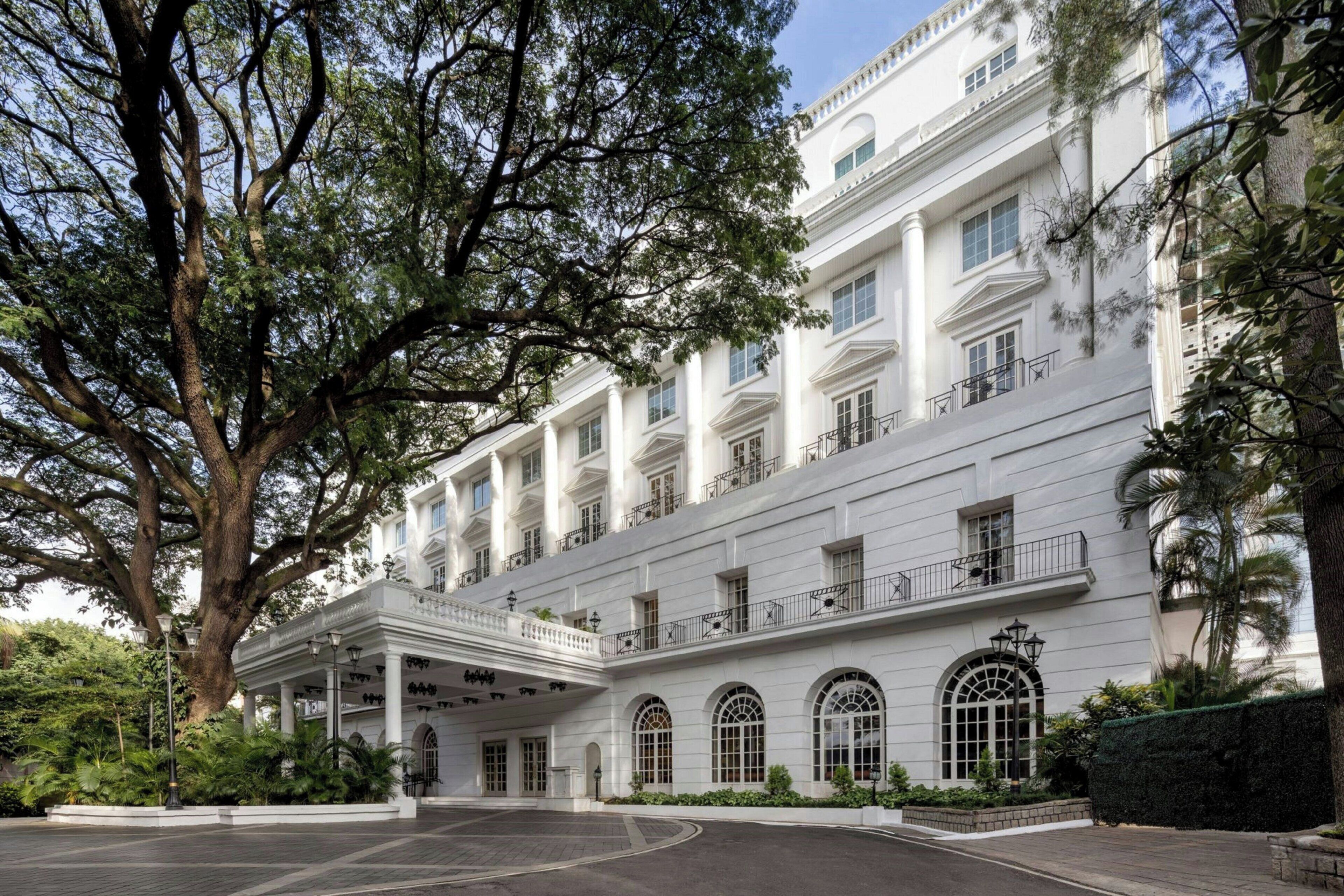 Un edificio blanco y majestuoso con un vestíbulo cubierto, ventanas amplias y un árbol en primer plano.