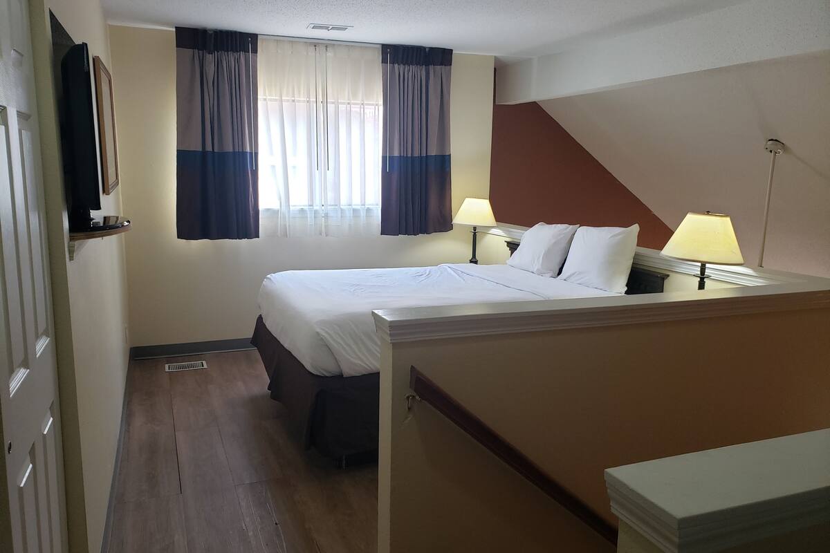 Une chambre d’hôtel avec un lit, des lampes de chevet, une fenêtre avec des rideaux, un téléviseur fixé au mur et un plancher en bois.
