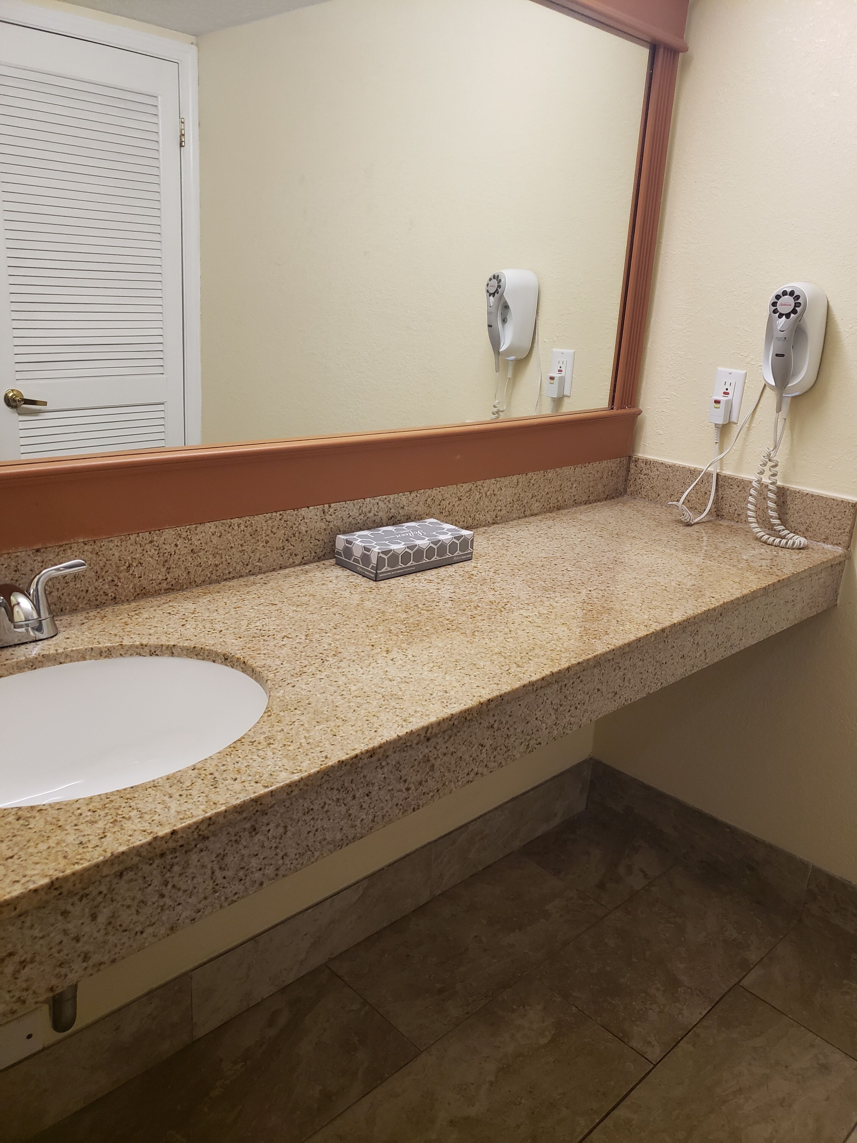 Un baño con encimera de granito, un lavamanos blanco, un espejo y dos secadores de pelo fijados a la pared.