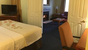 A hotel room with a bed, a TV, a fireplace, a chair, and a table. - Birch Hill Grand Rapids (Grand Rapids)