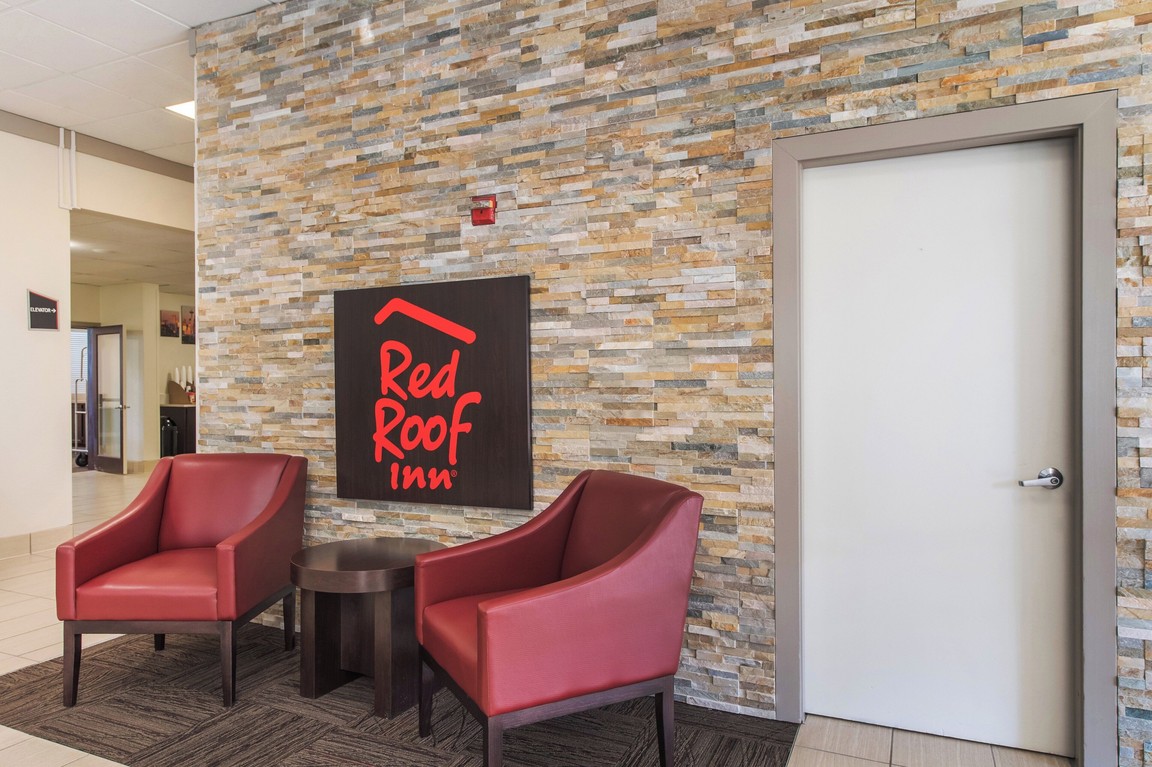 Señalización de Red Roof Inn sobre una pared de piedra texturizada, dos sillones de cuero rojo, una mesita redonda y una puerta.