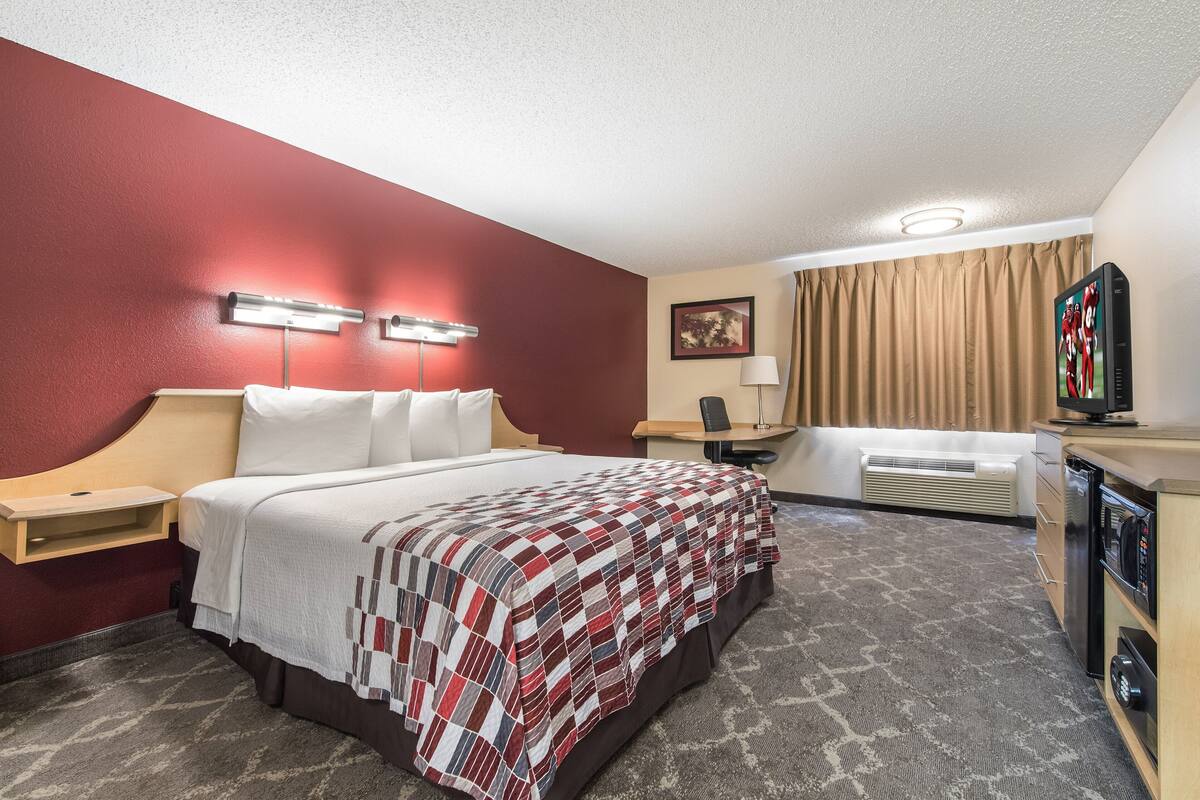 A hotel room with a bed, bedside tables, a desk, a chair, a television, and a minibar.
