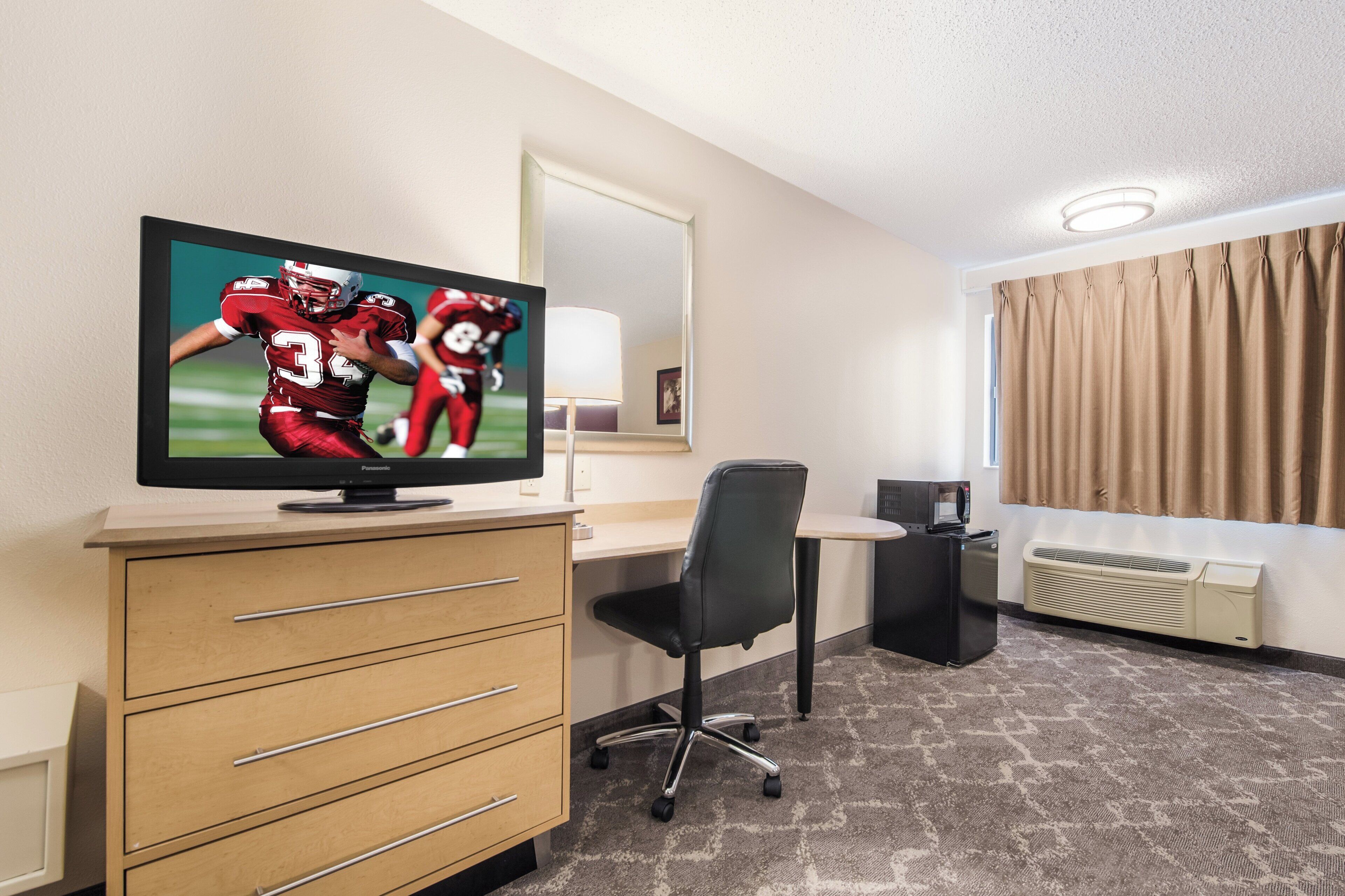 A hotel room with a flat-screen TV, a desk, a chair, a dresser, a mirror, a radiator, and a window with curtains.