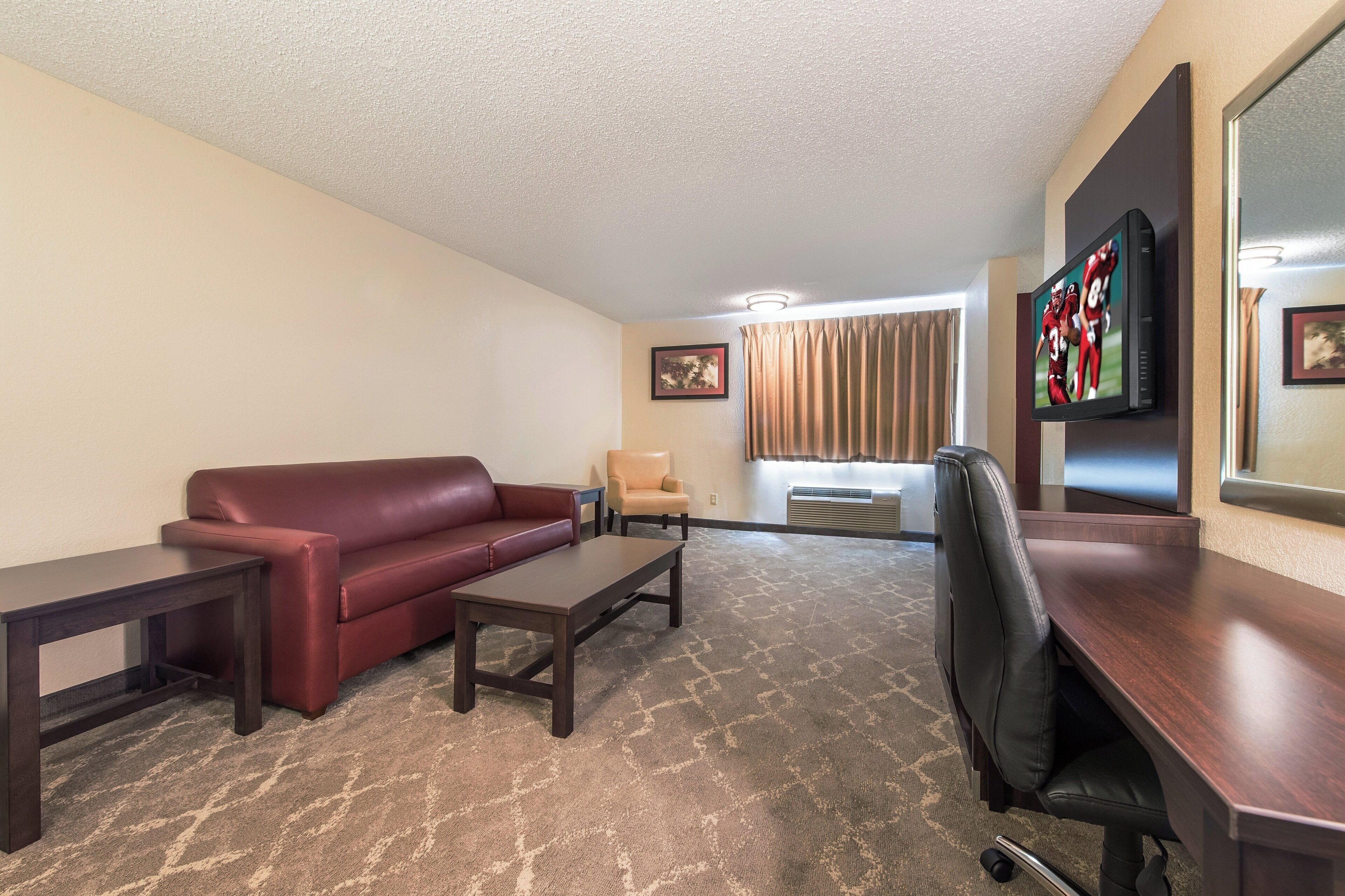 A hotel room with a leather sofa, a small table, a desk, a chair, a television, and a window with curtains.