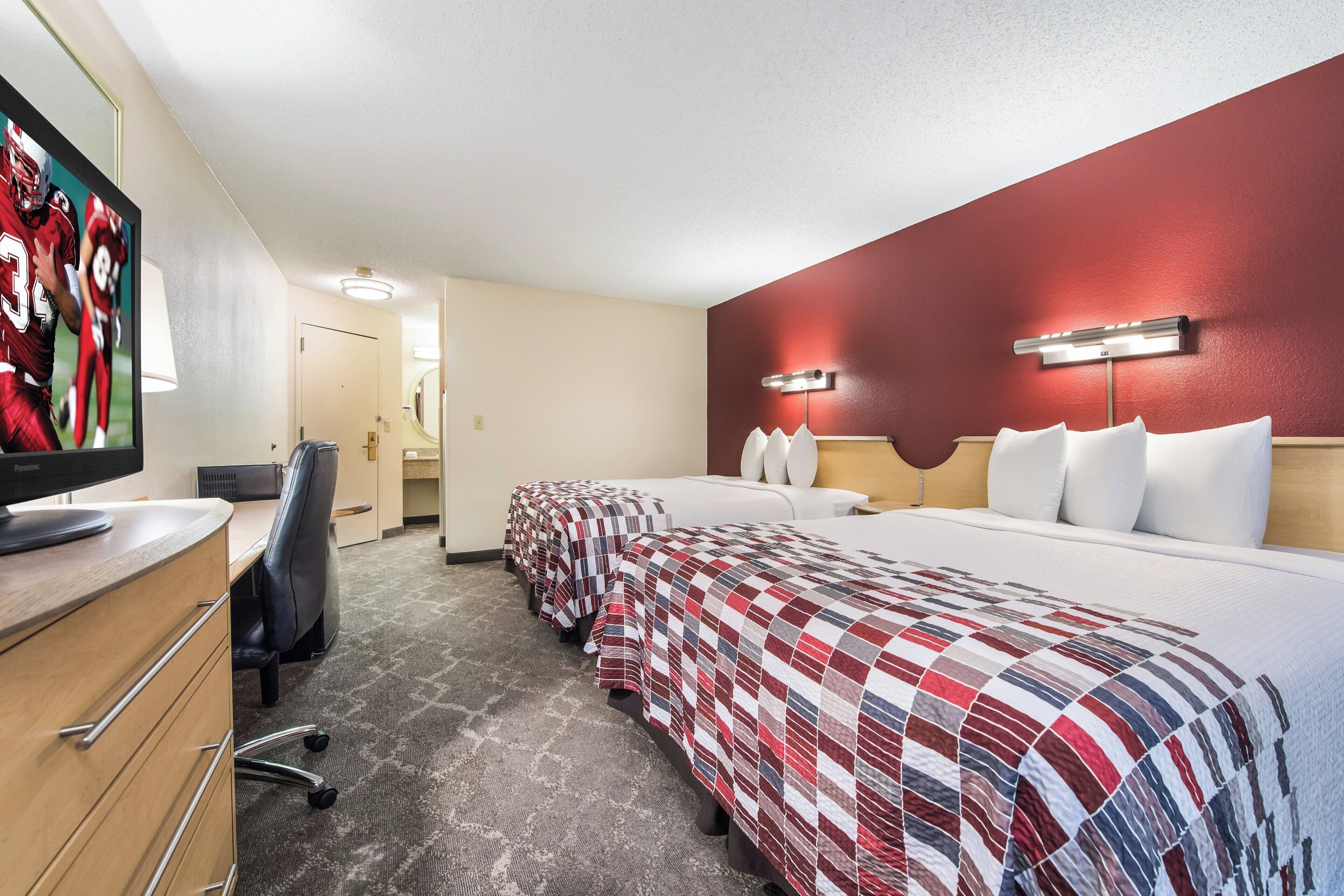 A hotel room with a television, a desk, a chair, a bed with a red and white quilt, and a red accent wall.