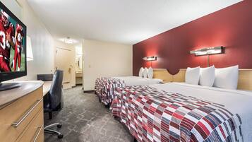 A hotel room with a television, a desk, a chair, a bed with a red and white quilt, and a red accent wall.