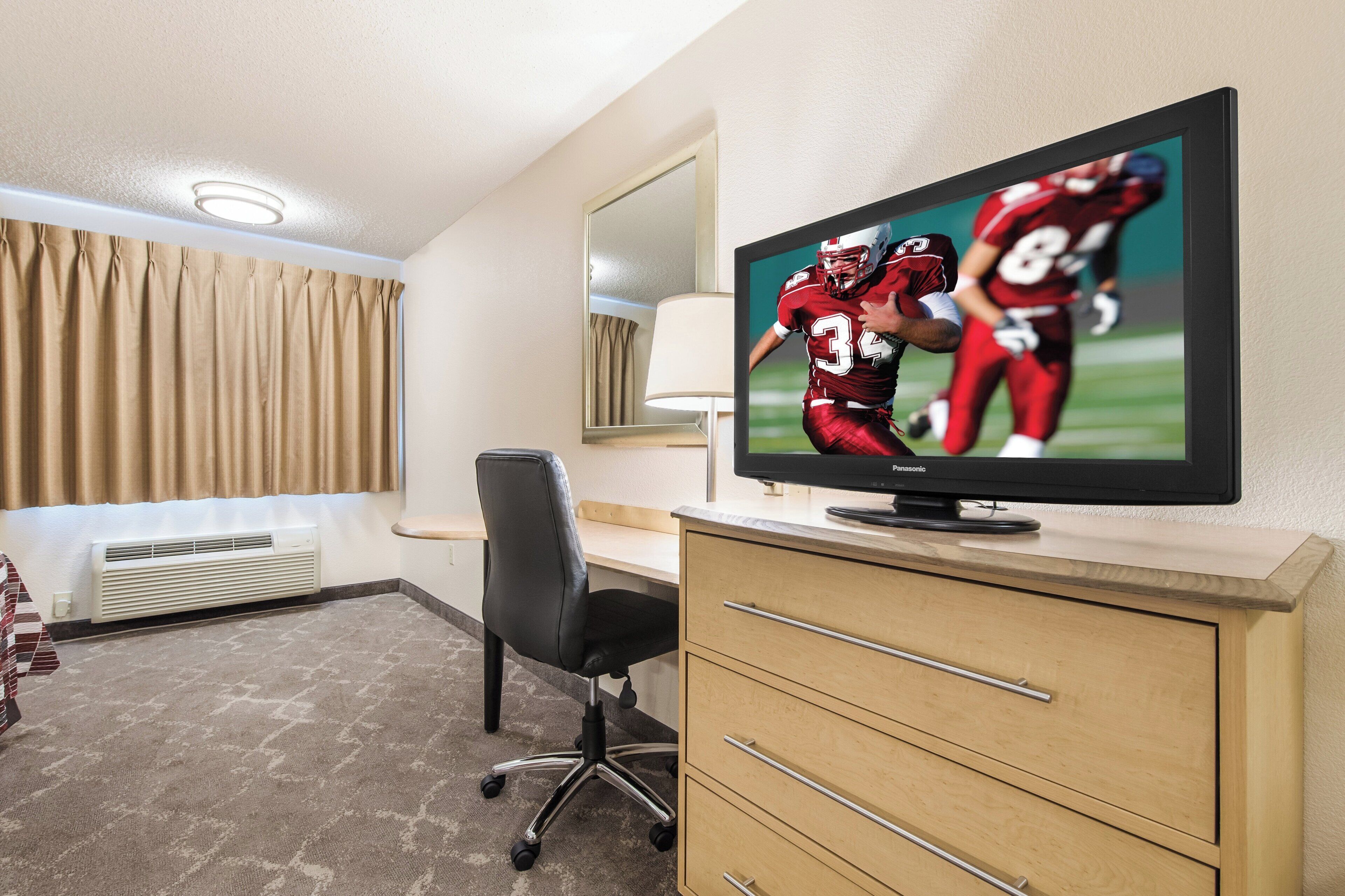 Habitación de hotel con una televisión mostrando un partido deportivo, un escritorio, una silla, una lámpara y una ventana con cortinas.