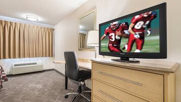 Habitación de hotel con una televisión mostrando un partido deportivo, un escritorio, una silla, una lámpara y una ventana con cortinas.
