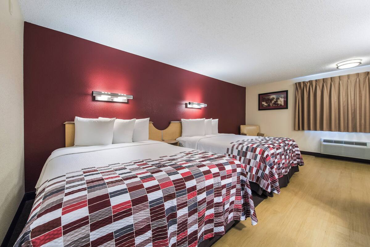 A hotel room with two beds, each with a red and white checkered bedspread, a red accent wall, and a picture on the wall.