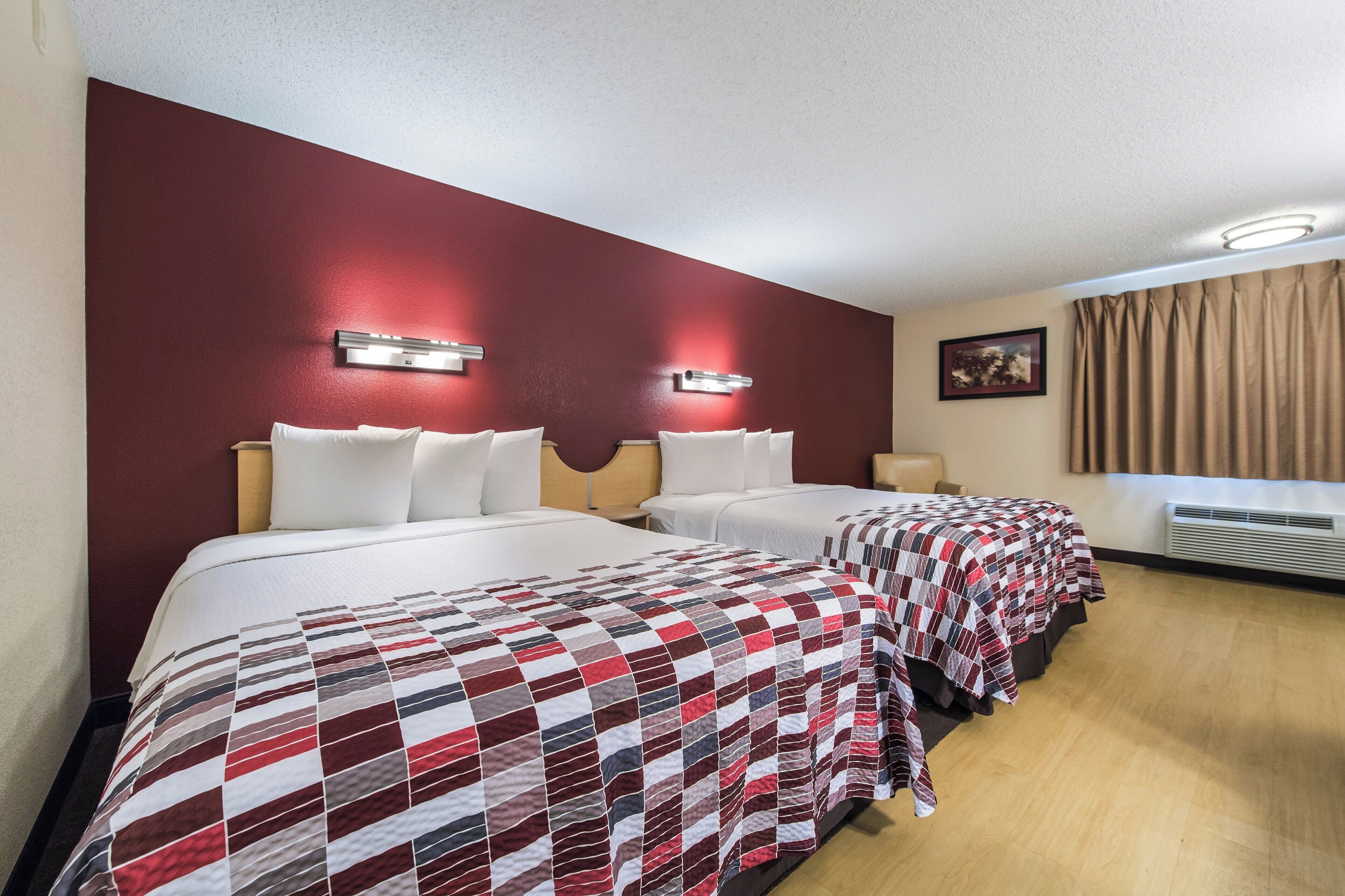 Ein Hotelzimmer mit zwei Betten, einer roten Wand als Akzent, einem Gemälde und einem Fenster mit Vorhängen.