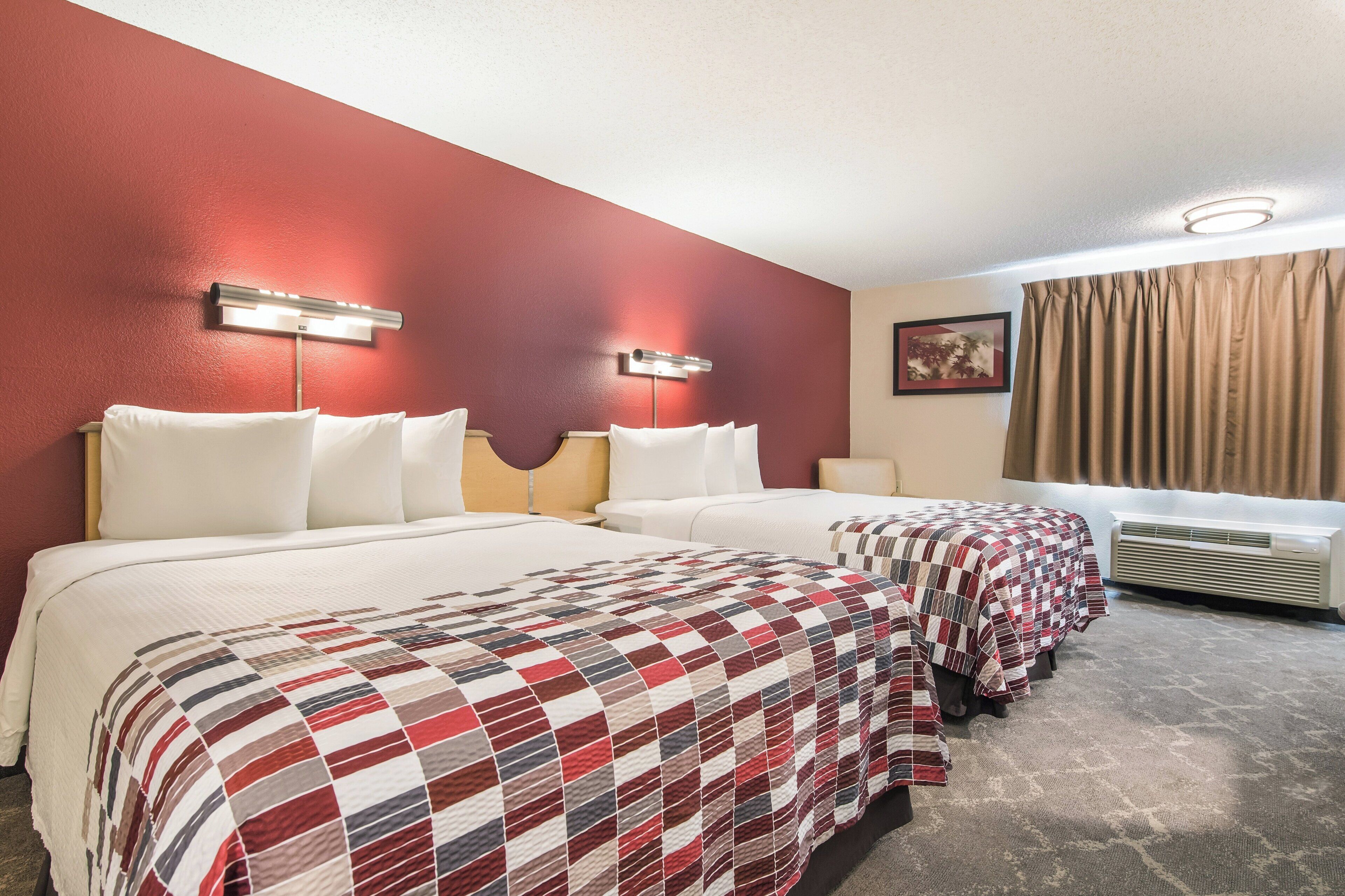 A hotel room with two beds, a window with curtains, an air conditioning unit, and a wall-mounted light fixture.