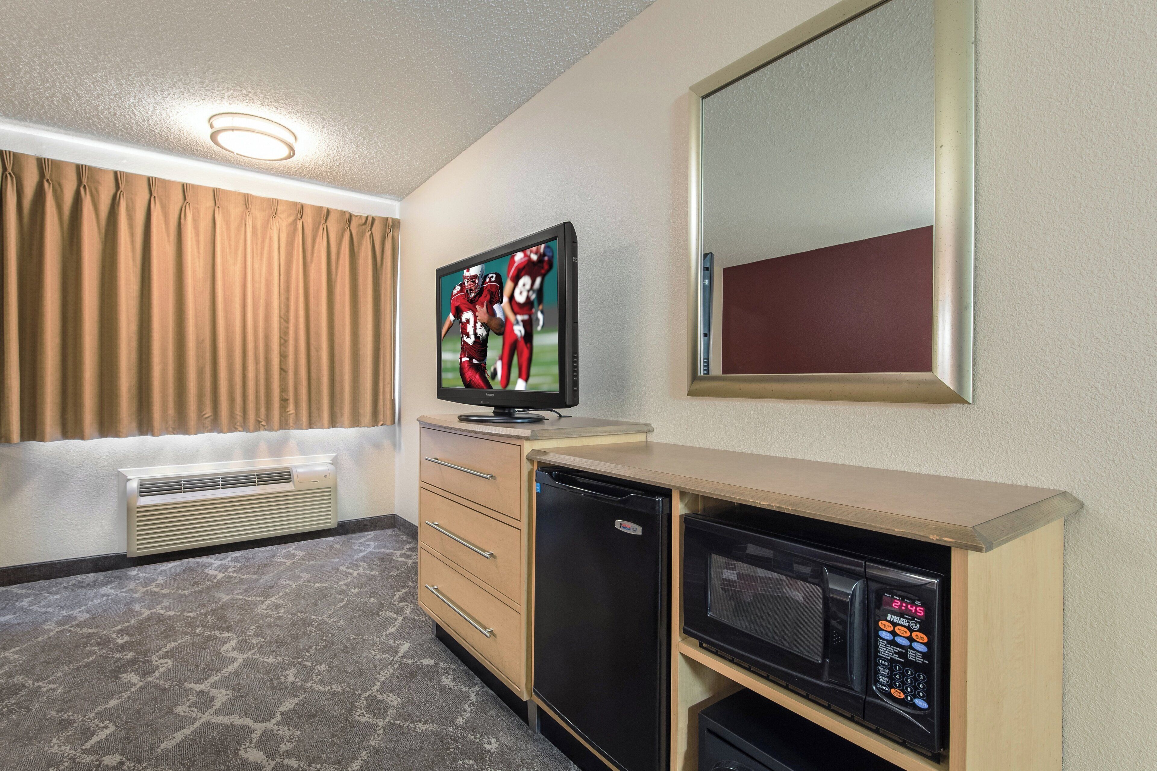 A hotel room with a flat-screen TV, a small refrigerator, a microwave, a wooden cabinet, a heater, and a window with curtains.