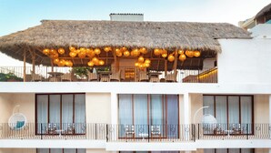 Un hotel de varios pisos con balcones, mobiliario exterior y un techo de paja en la planta superior.
