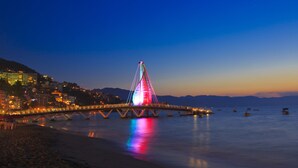 Un puente con luces de colores al anochecer, una playa tranquila y un paisaje urbano de fondo.