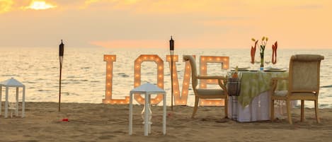 A beachside dining setup with a table, chairs, and a view of the ocean at sunset.
