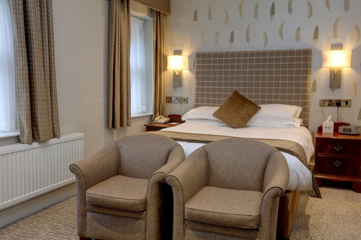 Une chambre d’hôtel avec un lit, deux fauteuils, une table de chevet, une lampe et une fenêtre avec des rideaux.