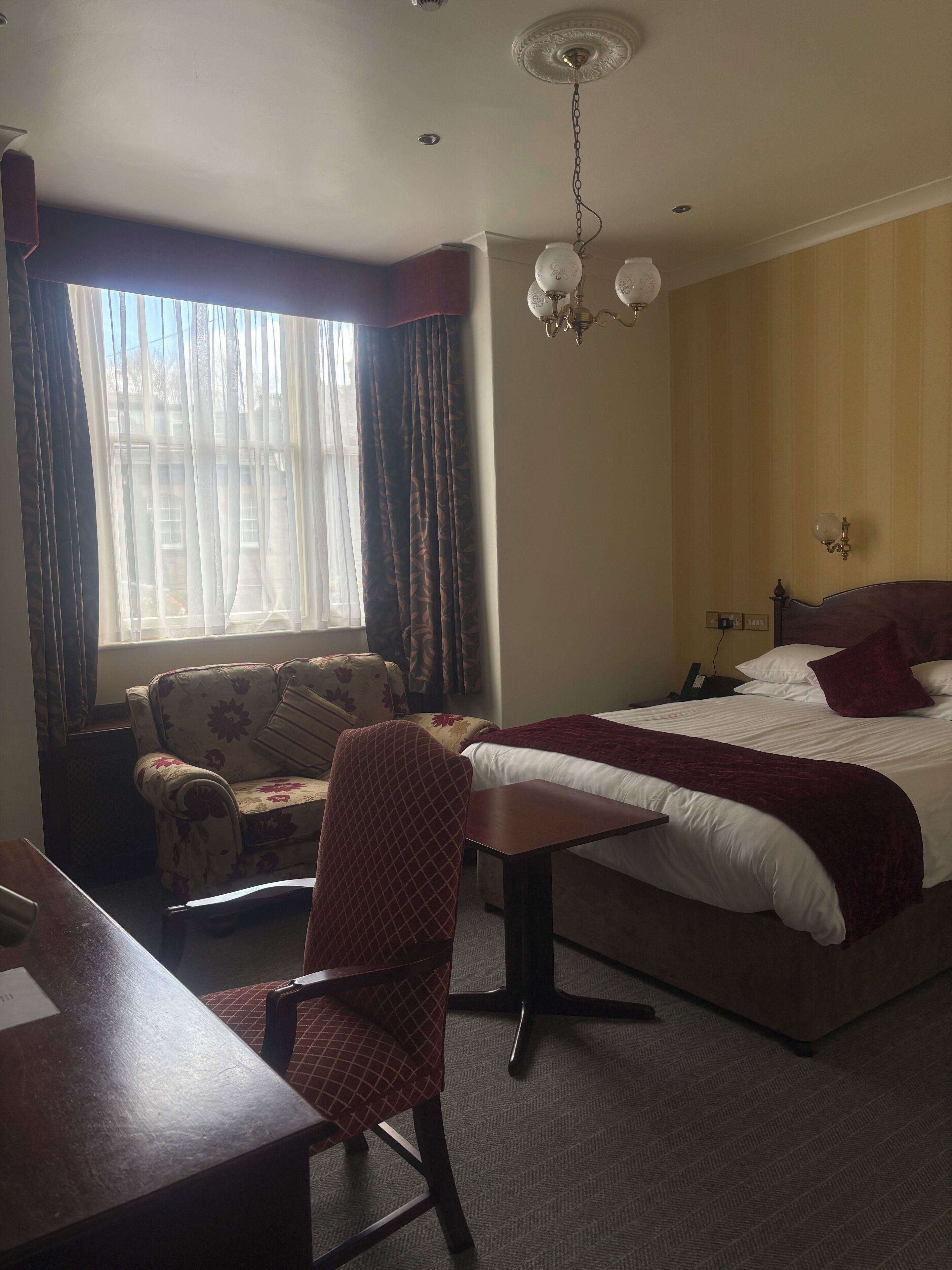 Una habitación de hotel con cama, escritorio, silla, candelabro y ventana con cortinas.