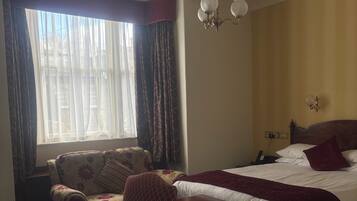 Una habitación de hotel con cama, escritorio, silla, candelabro y ventana con cortinas.