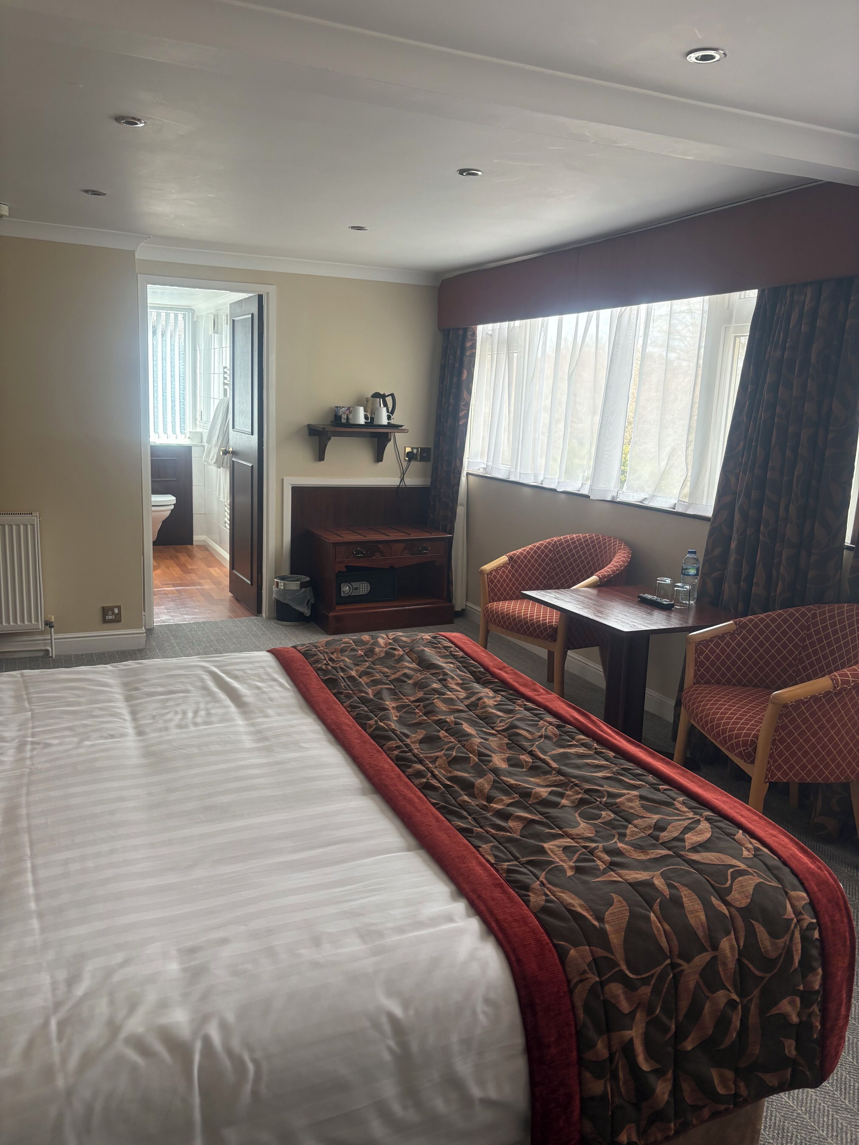 A hotel room with a bed, a desk, two chairs, a window with curtains, a shelf with items, and a view into another room.