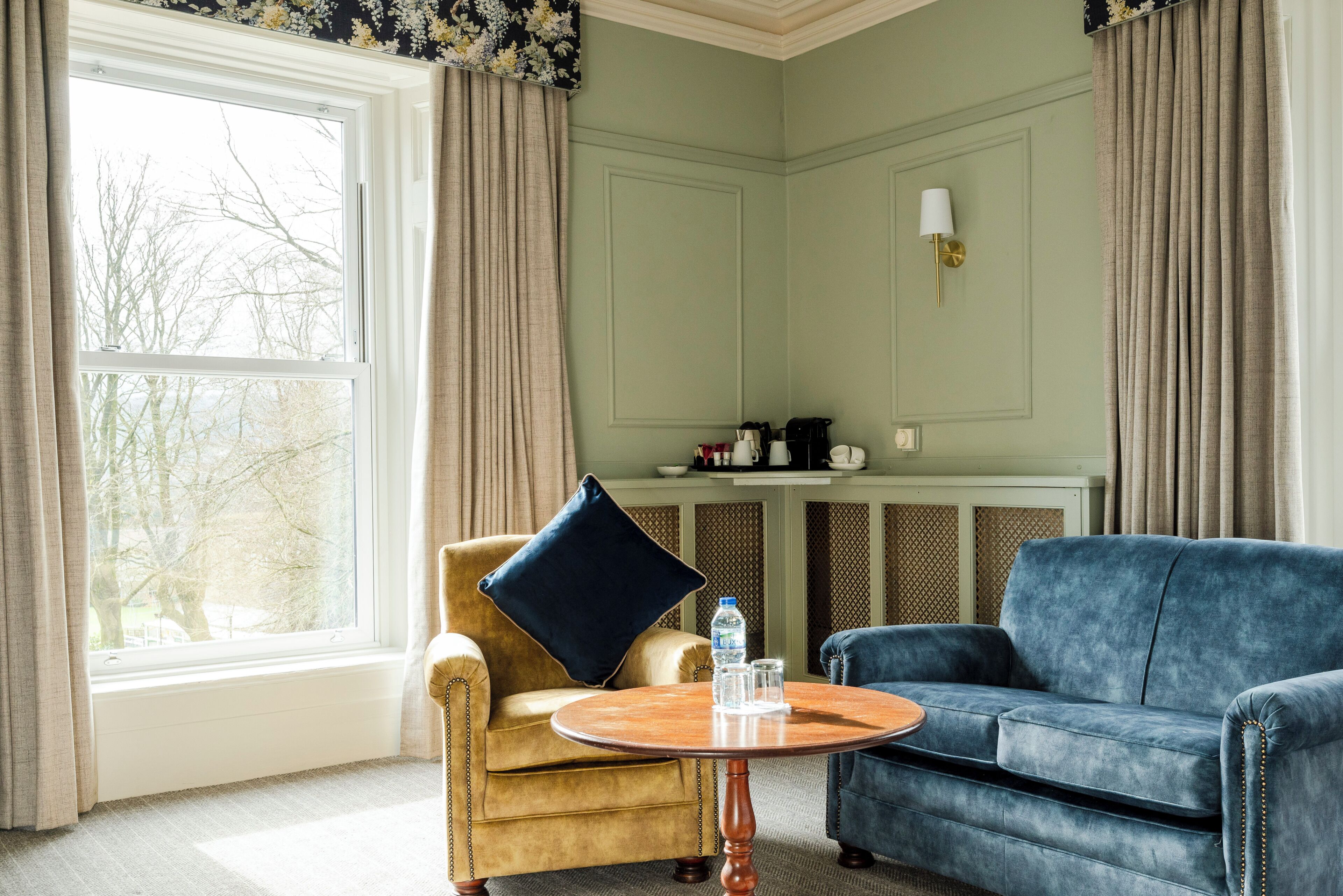 A room with a large window, a round wooden table, a blue sofa, and a gold-armored armchair.