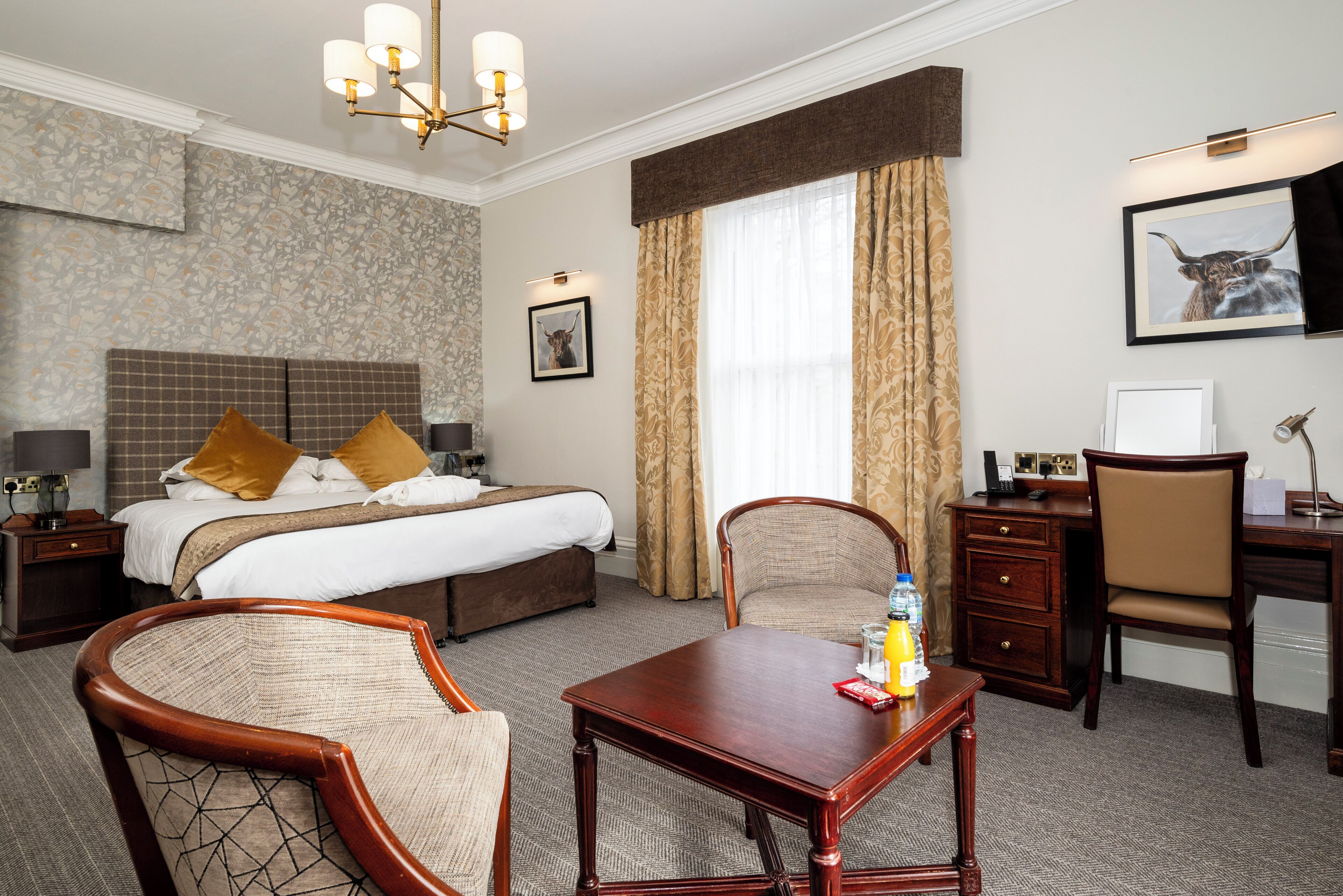 A hotel room with a bed, two chairs, a desk, a lamp, a picture on the wall, and a window with curtains.