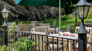 An outdoor dining area with wooden tables and chairs, covered by a green canopy, and surrounded by a garden with flowering bushes.