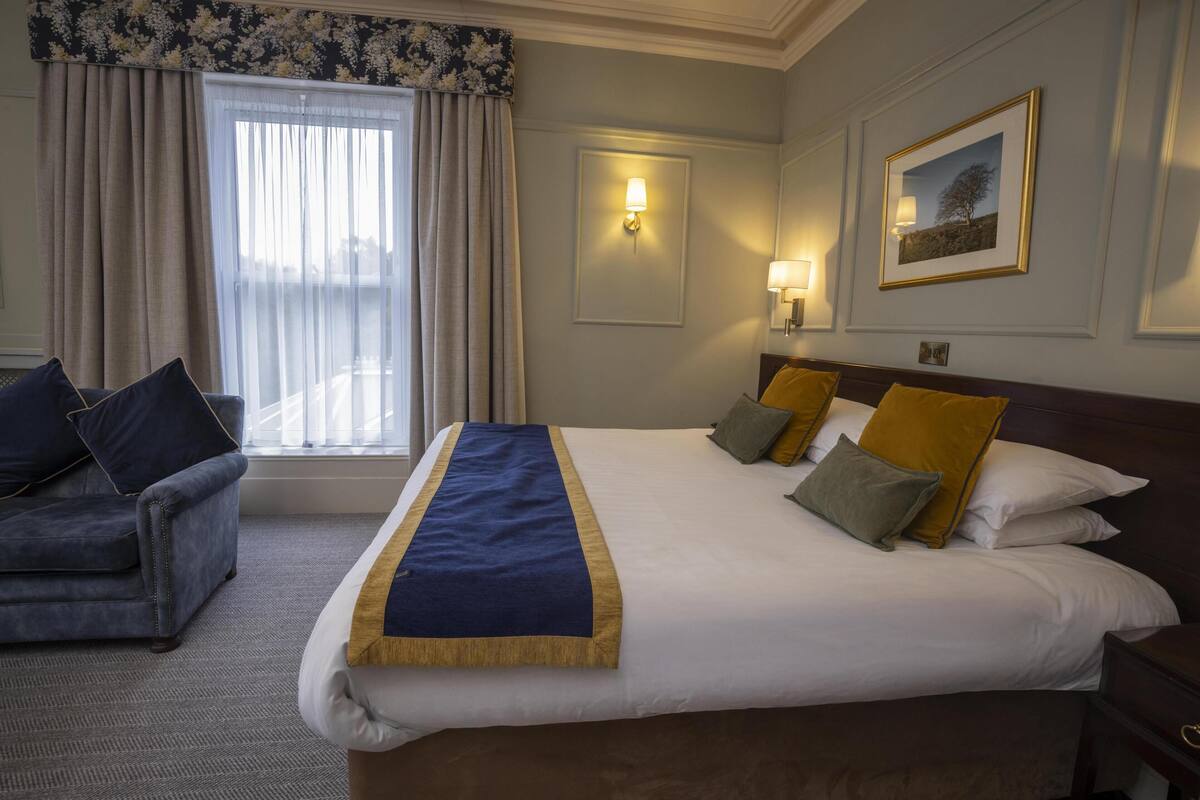 Une chambre d’hôtel avec un grand lit, un canapé, une fenêtre avec des rideaux, une lampe fixée au mur et un tableau encadré.