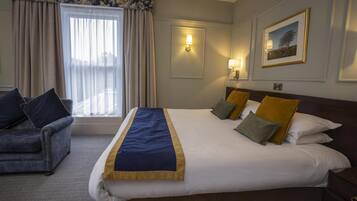 A hotel room with a large bed, a sofa, a window with curtains, a wall-mounted lamp, and a framed picture.