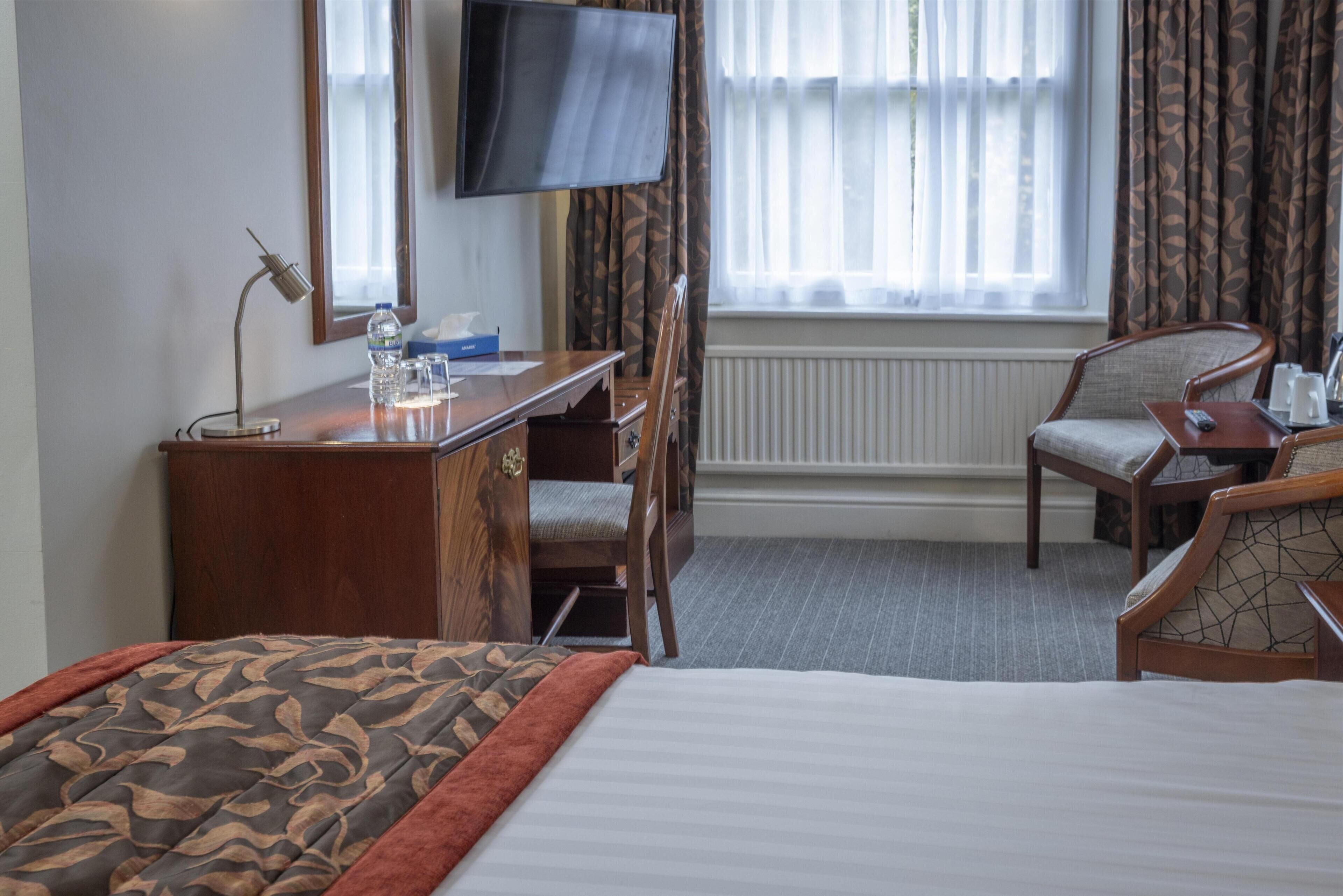 Quarto de hotel com cama, escrivaninha, cadeira, televisão e janela com cortinas.