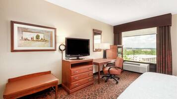 A hotel room with a bed, a desk with a TV, a chair, a dresser, and a large window with a view of buildings.